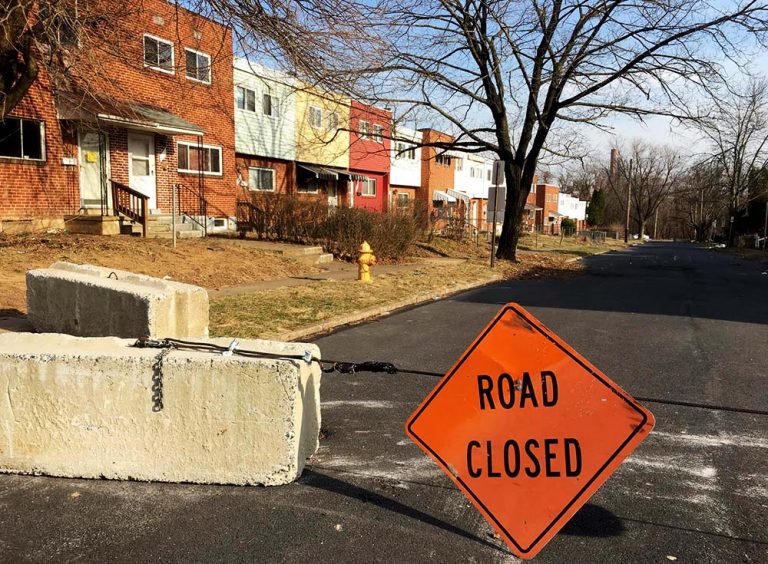 orange road closed sign hanging in middle of street. Can see colorful, newer row homes.