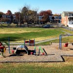 green grass in bckground, playground in foreground. Blue sky, rowhomes behind it.