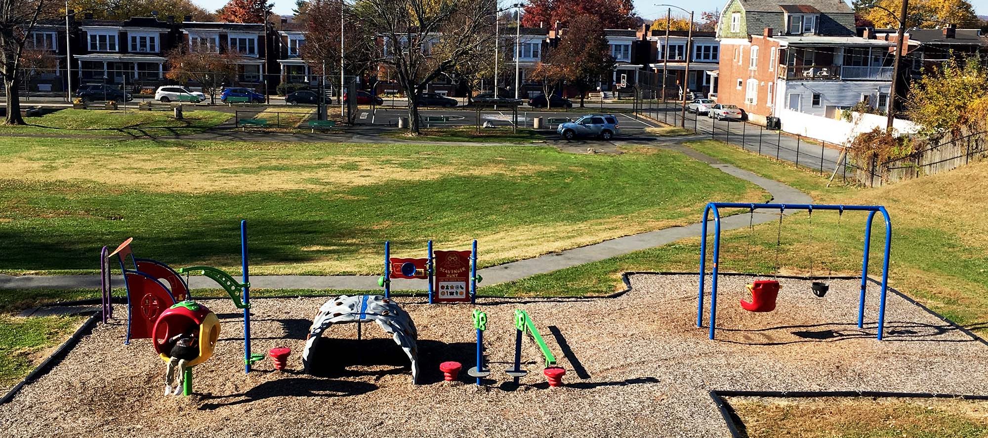 green grass in bckground, playground in foreground. Blue sky, rowhomes behind it.