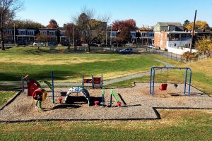 green grass in bckground, playground in foreground. Blue sky, rowhomes behind it. 