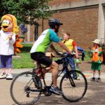 man with green and white bike shirt and helmet is riding a bike. Some kids play in background. Person wearing a turkey costume stand behind.