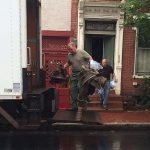 two white men loading a truck with what appear to be old chairs. They stand outside a red brick home